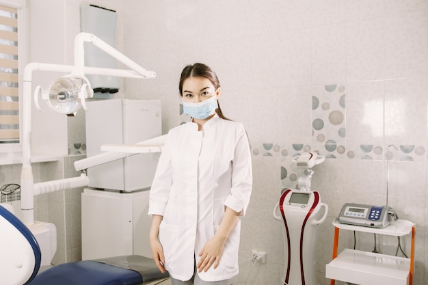 Dentista femminile allegro che sorride al suo ufficio. La bella studentessa in una maschera protettiva in odontoiatria sta nell'ufficio dentale