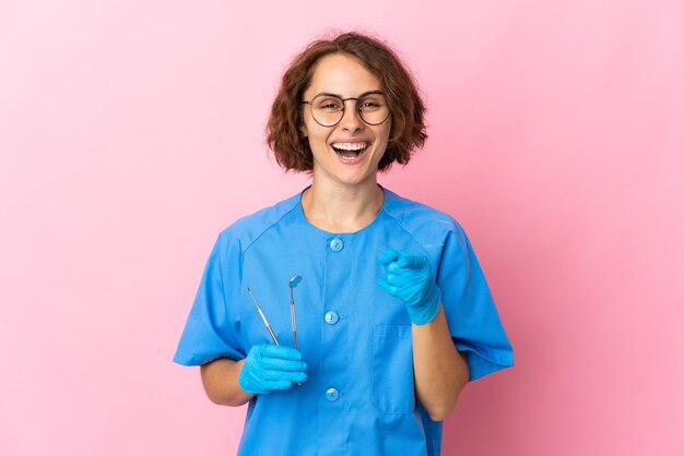 Dentista inglese della donna che tiene gli strumenti sopra isolato sulla parete rosa sorpreso e che indica la parte anteriore