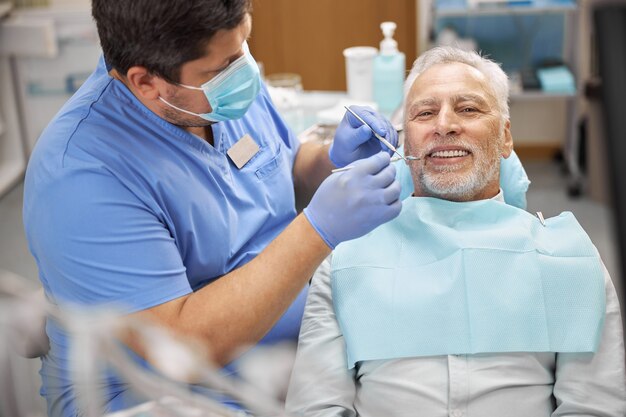 Dentista maschio qualificato che esamina i denti di un uomo anziano seduto su una poltrona del dentista