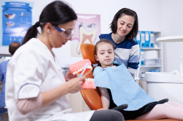 Dentista pediatrico che educa la bambina a lavarsi i denti nel modo corretto. Stomatolog d'ascolto della bambina e della madre che parlano dell'igiene dei denti nella clinica di odontoiatria che tiene il modello della mandibola.