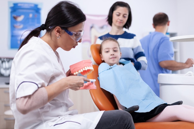 Dentista pediatrico che tiene il modello della mascella che spiega la cavità al bambino che indossa la pettorina. Stomatolog d'ascolto della bambina e della madre che parlano dell'igiene dei denti nella clinica di odontoiatria che tiene il modello della mandibola.