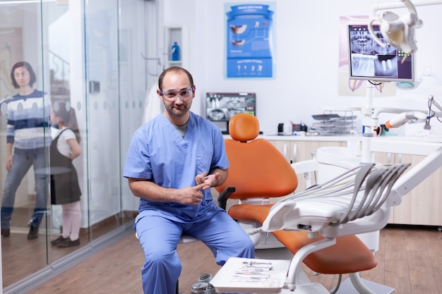 Dentista professionista che discute di più problemi di igiene dei denti seduto su una sedia. Stomatolog nella clinica dei denti professioanl sorridente indossando l'uniforme che guarda l'obbiettivo.