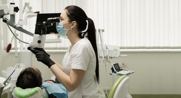 Dentista professionista della donna che esegue varie procedure odontoiatriche e protesiche a un paziente in un ufficio moderno utilizzando una vista laterale del microscopio