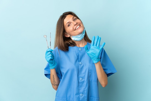 Dentista slovacco che tiene gli strumenti isolati sulla parete blu che saluta con la mano con l'espressione felice