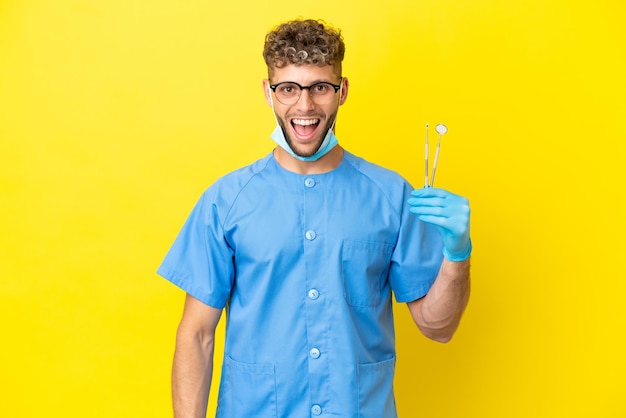 Dentista uomo biondo che tiene strumenti isolati su sfondo con espressione facciale a sorpresa