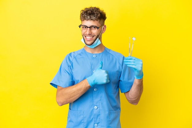 Dentista uomo biondo che tiene strumenti isolati su sfondo dando un pollice in alto gesto