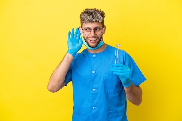 Dentista uomo caucasico tenendo gli strumenti isolati su sfondo giallo ascoltando qualcosa mettendo la mano sull'orecchio