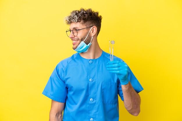 Dentista uomo caucasico tenendo gli strumenti isolati su sfondo giallo guardando lato