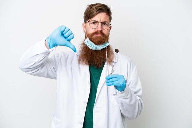 Dentista uomo rossastro che tiene strumenti isolati su sfondo bianco che mostra il pollice verso il basso con espressione negativa