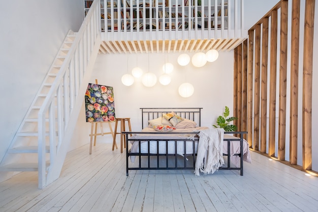 Design moderno di lusso di un accogliente monolocale in stile scandinavo con pareti bianche, secondo piano con una biblioteca e un'enorme finestra alta piena di luce del giorno