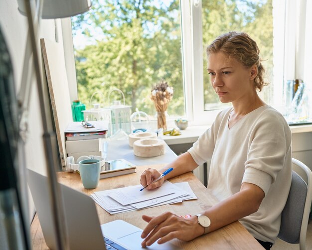 Designer di giovane imprenditrice femminile che scrive sul foglio di carta, guardando il laptop, pensiero serio della donna