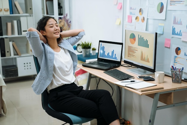 Designer femminile creativo asiatico che si tiene per mano dietro e chiude l'occhio davanti al computer sulla scrivania. giovane ragazza dipendente rilassarsi dal duro lavoro in un moderno ufficio luminoso. la donna sorridente gode dello stretching della pausa