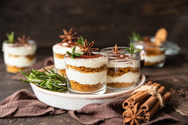 Dessert a strati in vasetto di vetro con crumble di biscotti e panna montata decorato con rosmarino e anice, superficie marrone scuro