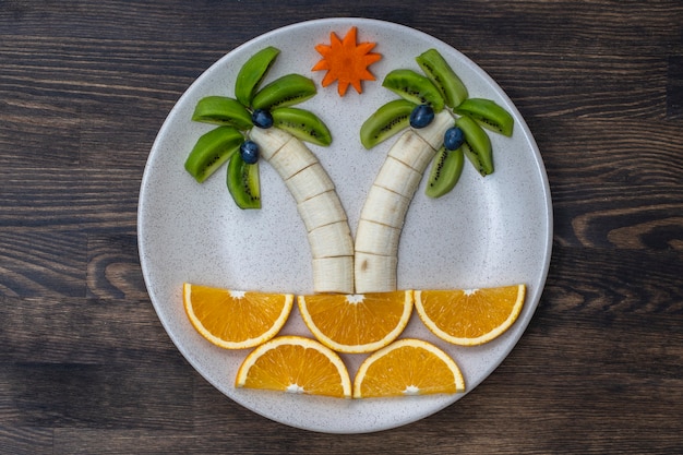 Dessert alla frutta creativo con kiwi, banana, uva, carota e arancia. Concetto di cibo per bambini. Macedonia di frutta divertente e sana per i bambini. Forma di alberi di palma a base di frutta in zolla bianca sulla tavola di legno