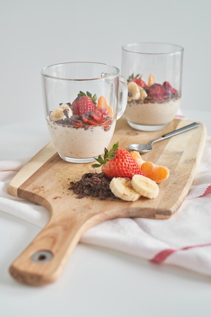 Dessert allo yogurt alla fragola con cioccolato grattugiato guarnito in una tazza di vetro e servito su tagliere di legno con cucchiaio e fette di mandarino e banana vicino al tovagliolo