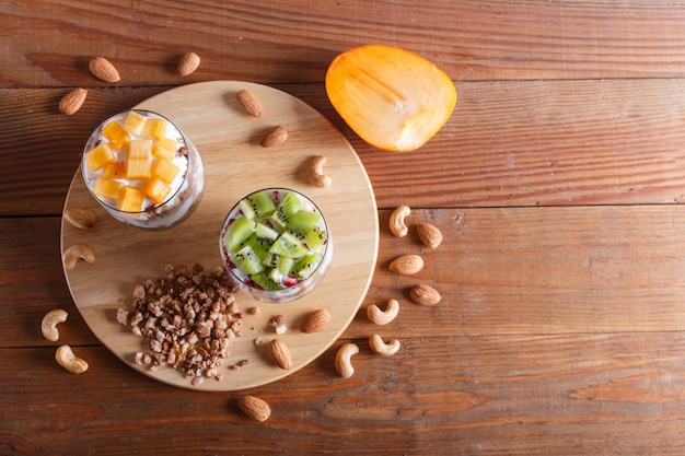 Dessert con yogurt greco, granola, mandorle, anacardi, kiwi e cachi su fondo di legno marrone.