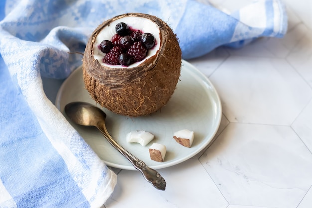 Dessert della bacca con yogurt in una tazza della noce di cocco su un fondo leggero. More, lamponi, mirtilli, ribes nero. Tovagliolo blu