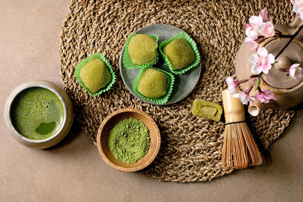 Dessert di riso asiatico dolce verde matcha mochi con tazza di tè matcha schiumato e polvere di matcha verde in piatto di ceramica sul tavolo marrone Teiera e ramo fiorito rosa primaverile su tovagliolo di paglia lavorato a maglia