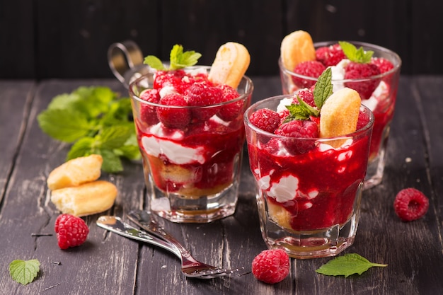 Dessert italiano tradizionale - dolce di tiramisu con il lampone e menta in vetri sul fondo di legno del nero di lerciume.