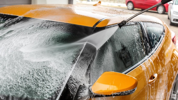 Dettagli sul parabrezza dell'auto giallo scuro e sullo specchietto laterale che viene lavato con uno spray a getto d'acqua, schiuma di sapone per la pulizia.