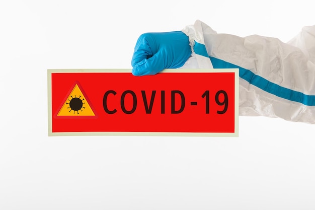 Dettaglio delle braccia di un'infermiera medica che indossa un DPI e guanti in lattice con un cartello rosso che recita: "COVID-19". Coronavirus, pandemia e concetto di salute.