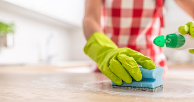 Dettaglio di una mano in un guanto di gomma e una spugna blu che puliscono una superficie con un detergente.