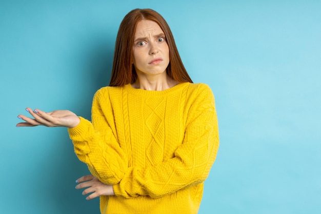 Di che diavolo stai parlando. Studio shot di dispiaciuta donna confusa con i capelli rossi che gesticolano con il palmo alzato, accigliata su sfondo blu. Concetto di emozioni negative.