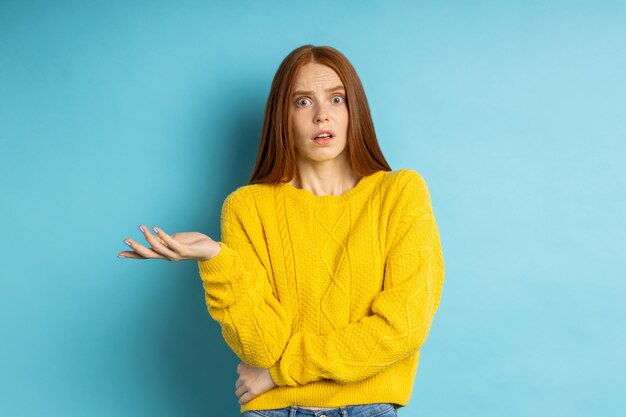 Di che diavolo stai parlando. Studio shot di dispiaciuta donna confusa con i capelli rossi che gesticolano con il palmo alzato, accigliata su sfondo blu. Concetto di emozioni negative.