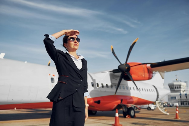 Di giorno la giovane hostess che indossa abiti neri formali è in piedi all'aperto vicino all'aereo