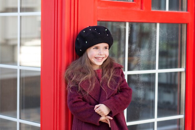 Di nuovo al concetto di scuola, autunno. La bambina allegra si trova vicino alla cabina telefonica rossa di Londra in un bellissimo cappotto bordeaux e prende e sorride. Inghilterra, Regno Unito Viaggio in Europa. Formazione scolastica.