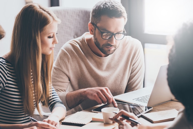 Diamo un'occhiata alle nostre statistiche! Giovane uomo bello che indica la tavoletta digitale mentre è seduto al tavolo dell'ufficio per un incontro di lavoro con i suoi colleghi