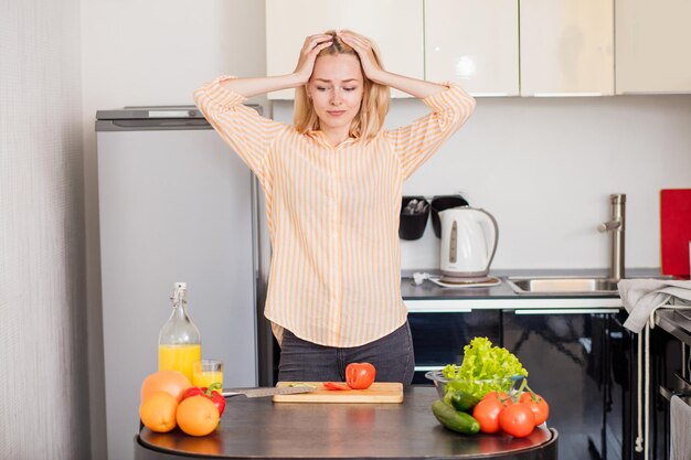 Dieta, perdita di peso, concetto di cucina. Signora bionda caucasica confusa perplessa in piedi accanto al tavolo con frutta e verdura in cucina, toccando la testa con entrambe le mani.