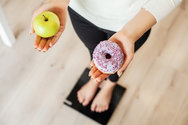 Dieta. Peso corporeo di misurazione della donna sulla ciambella e sulla mela della tenuta della bilancia. I dolci sono cibo spazzatura malsano. Fast food