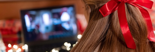 Dietro la testa femminile lunghi capelli biondi e fiocco rosso vista posteriore guardando il film di Natale sul computer portatile in campeggio a casa in un letto accogliente con ghirlanda di luci incandescenti di notte a natale o capodanno banner