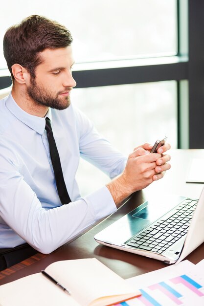 Digitando un messaggio aziendale. Vista dall'alto del giovane in camicia e cravatta che tiene il telefono cellulare e lo guarda mentre è seduto al suo posto di lavoro