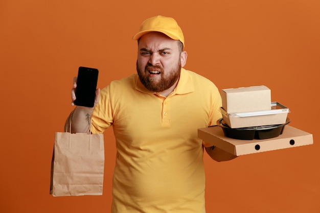 Dipendente dell'uomo delle consegne in uniforme della maglietta vuota con cappuccio giallo che tiene contenitore per alimenti e scatola per pizza con sacchetto di carta che mostra lo smartphone con la faccia arrabbiata in piedi su sfondo arancione