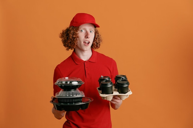 Dipendente dell'uomo delle consegne in uniforme della maglietta vuota con cappuccio rosso che tiene contenitori per alimenti e tazze di caffè guardando la fotocamera stupito e sorpreso in piedi su sfondo arancione