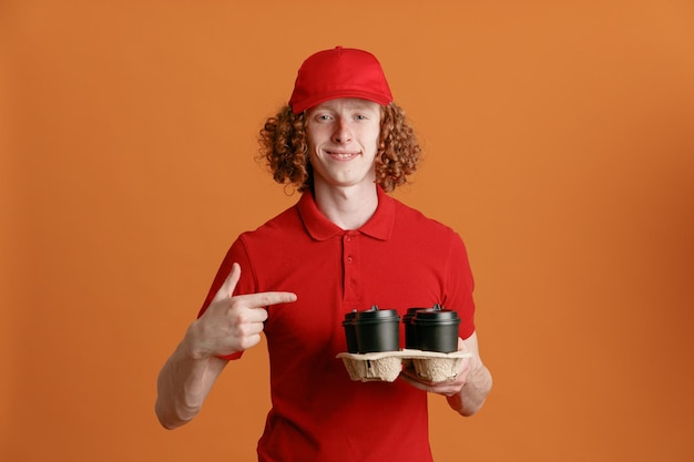 Dipendente dell'uomo delle consegne in uniforme della maglietta vuota con cappuccio rosso che tiene le tazze di caffè che puntano con il dito indice verso di loro sorridendo fiducioso in piedi su sfondo arancione