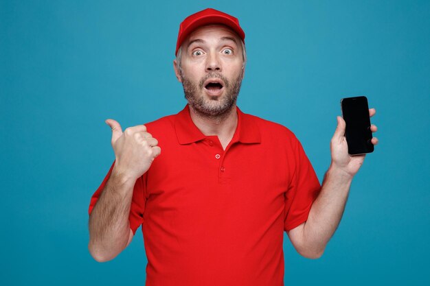 Dipendente dell'uomo delle consegne in uniforme della maglietta vuota con cappuccio rosso che tiene lo smartphone puntato con il pollice di lato guardando la fotocamera stupito e sorpreso in piedi su sfondo blu