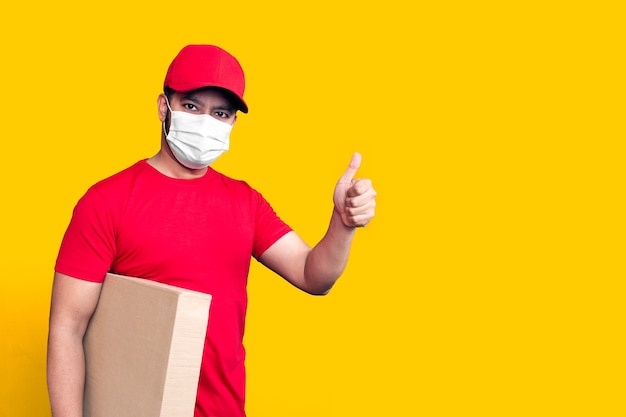 Dipendente dell'uomo di consegna in maschera facciale uniforme della maglietta in bianco del cappuccio rosso tiene la scatola di cartone vuota isolata su fondo giallo