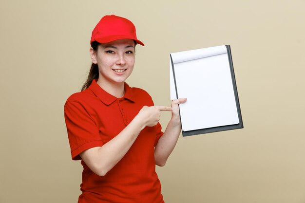 Dipendente di consegna donna in berretto rosso e maglietta vuota uniforme che tiene appunti e penna che punta con la penna agli appunti sorridendo allegramente in piedi su sfondo marrone