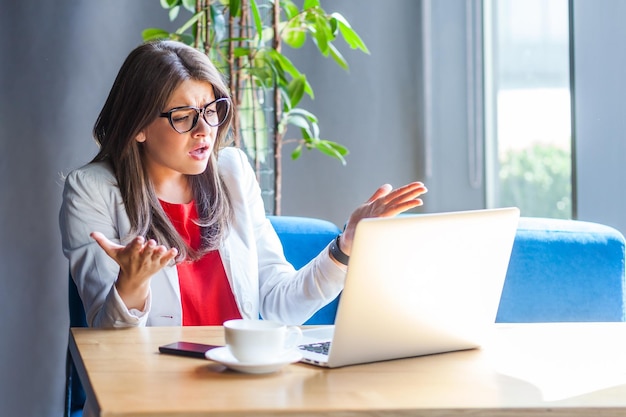 Dipendente donna infelice con gli occhiali che alza le mani in un gesto indignato chiedendo perché e guardando con rabbia guardando lo schermo del laptop parlando in videochiamata