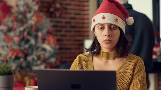 Dipendente femminile che analizza i dati di ricerca sul computer portatile, indossando il cappello di Babbo Natale durante le festività natalizie. Lavorare al computer per pianificare il rapporto prima di celebrare l'evento invernale. Scatto da treppiede.