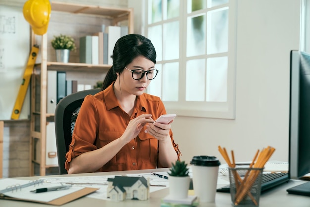 dipendente ingegnere donna asiatica seduto alla scrivania in ufficio controllando il progetto scatta foto inviando un messaggio tramite smartphone. amichevole piccola impresa donna architetto diagrammi e grafici di lavoro