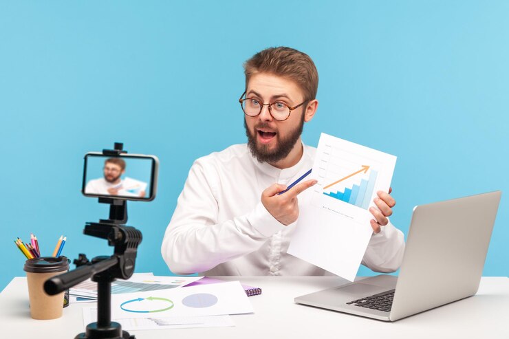 Dipendente maschio che analizza il grafico della crescita dei clienti registrando video sulla fotocamera dello smartphone o effettuando lo streaming online di conferenze che effettuano report Scatto in studio al coperto isolato su sfondo blu