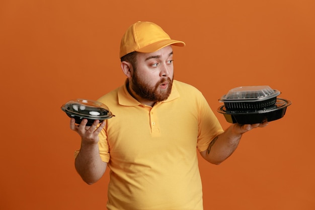 Dipendente uomo di consegna in uniforme tshirt bianca con cappuccio giallo che tiene contenitori per alimenti che sembrano confusi con dubbi in piedi su sfondo arancione