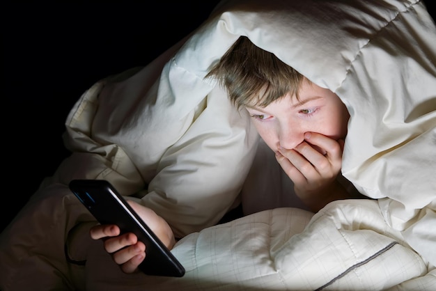 Dipendenza dai social media Il ragazzo che si nasconde sotto la coperta di notte nel suo letto comunica su Internet. Dipendenza da gadget per bambini e insonnia. ragazzo solitario che usa lo smartphone di notte a letto
