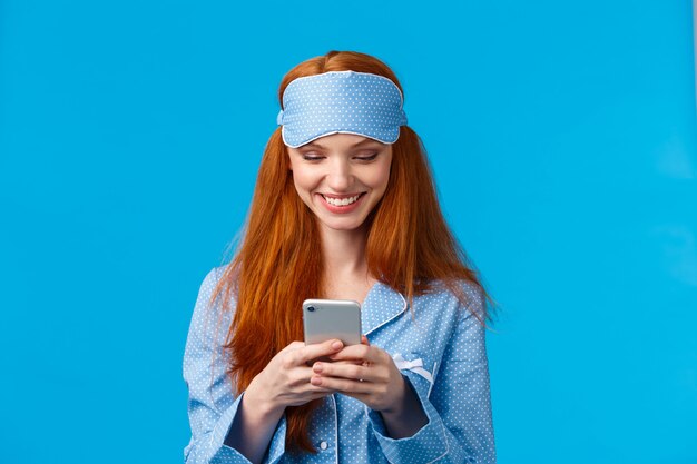 Dipendenza mobile, tecnologia e concetto di bellezza. Ragazza allegra che sveglia e prende il telefono, controllando i messaggi, scorrendo le notizie si nutrono di smartphone indossando indumenti da notte, maschera per dormire, sorridente