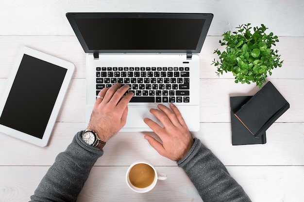 Direttamente sopra la vista delle mani umane che digitano sul laptop. Computer portatile, tablet digitale, diario, tazza di caffè e pianta in vaso sulla scrivania. Uomo che lavora da casa.