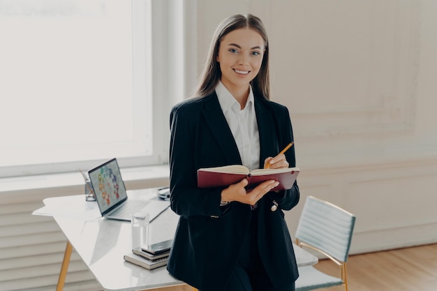 Direttore femminile che tiene taccuino e matita mentre si appoggia sulla scrivania dell'ufficio